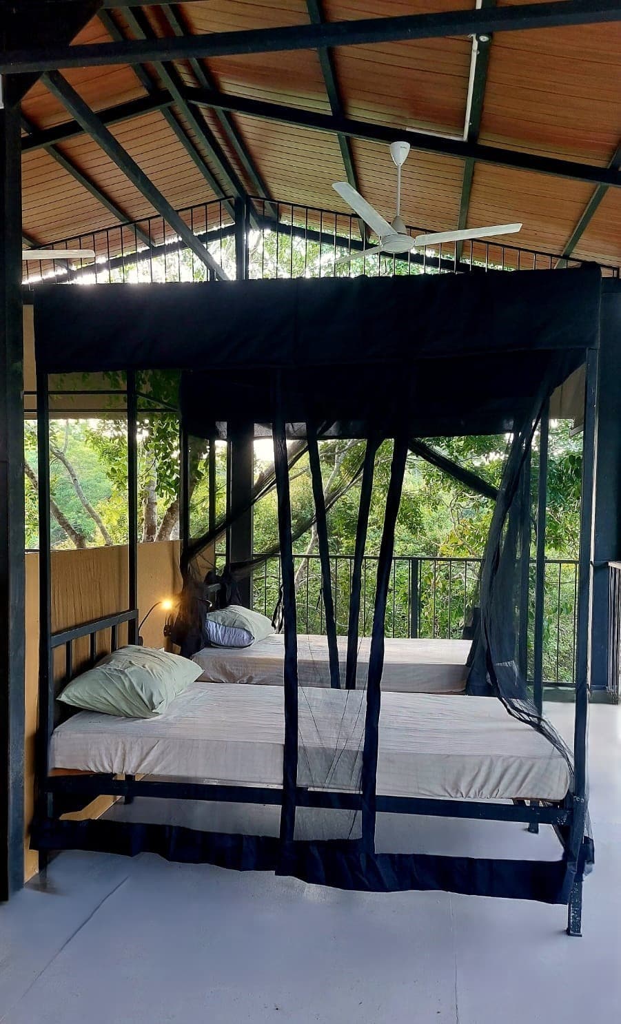 Two single beds under black mosquito nets on an open elevated platform, with a rail and green canopy directly beyond them