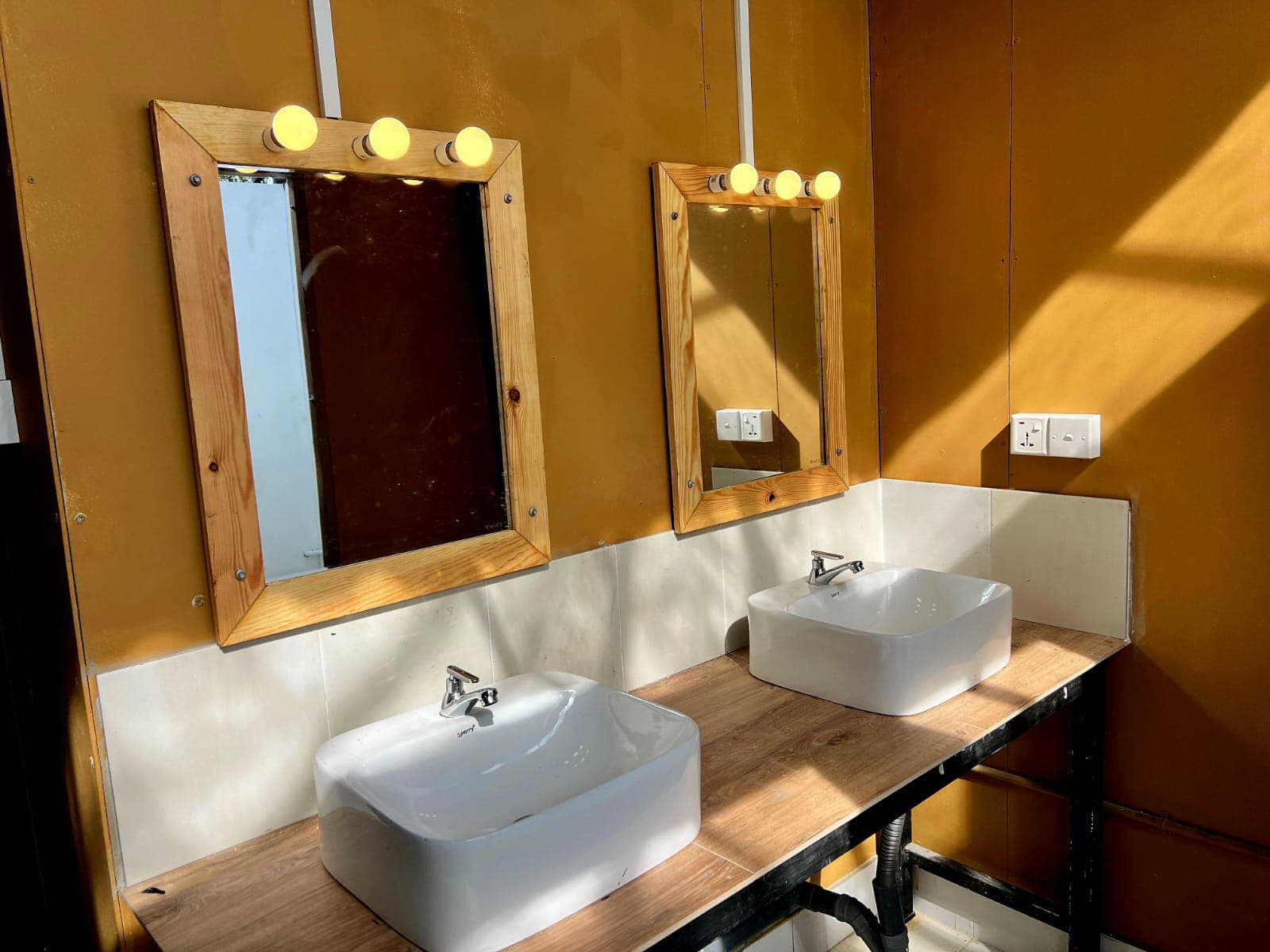 Two basins under bulb-lit mirrors against a deep yellow bathroom wall